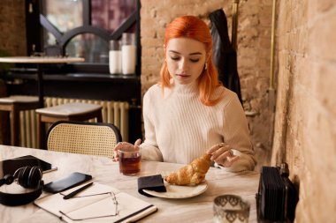 Modern bir kafede, kızıl saçlı genç bir kadın kruvasanla mola verir ve içer..