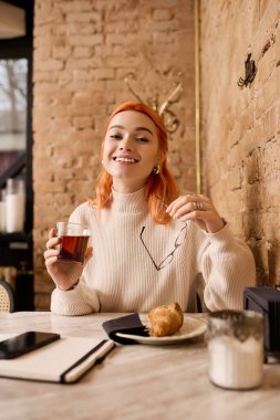Genç kızıl saçlı kadın, şık bir kafede çayın tadını çıkarırken neşeyle gülümsüyor..