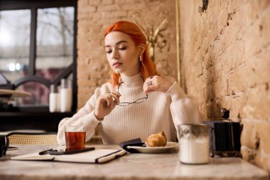 Kızıl saçlı genç bir kadın şık bir kafede dinleniyor, çay içiyor ve notlar yazıyor..