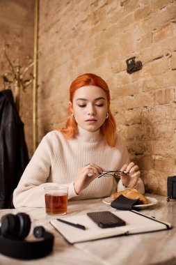 Şık bir kafede, kızıl saçlı genç bir kadın çayını yudumlarken ve pastanın tadını çıkarırken dinleniyor..