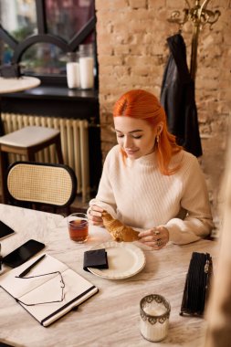 Canlı kızıl saçlı genç bir kadın şık bir kafede oturur, pastanın ve içkisinin tadını çıkarır..