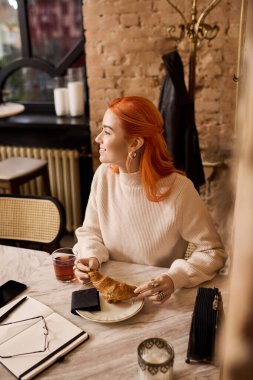 Kızıl saçlı güzel bir kadın, rahat bir kafede oturur, kruvasan ve çayın tadını çıkarır..