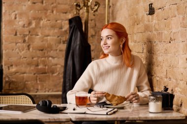 Kızıl saçlı neşeli genç bir kadın, sıcaklıkla çevrili, modaya uygun bir kafede pastanenin tadını çıkarıyor..