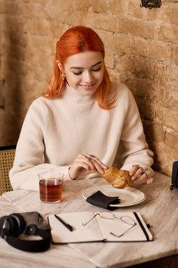 Kızıl saçlı genç bir kadın kruvasan ve çayın tadını çıkarırken gülümsüyor..
