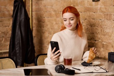 Canlı, kızıl saçlı genç bir kadın modern bir kafede gevşeyip, yiyecek ve içeceğinin tadını çıkarıyor..