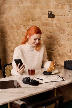 Kırmızı saçlı genç bir kadın kruvasan ve çay içerken şık bir kafede gülümsüyor..
