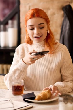 Kızıl saçlı neşeli genç bir kadın lezzetli bir kruvasanın tadını çıkarırken neşeli bir anı yakalıyor..