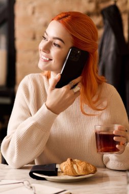 Moda bir kafede sıcak bir içecek yudumlarken kızıl saçlı neşeli genç bir kadın telefonunda sohbet ediyor..