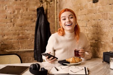 Kızıl saçlı neşeli genç bir kadın bir kafede oturur, içkisini yudumlar ve parlak bir şekilde gülümser..