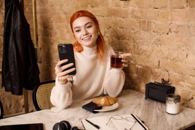 Güzel bir genç kadın elinde içki ve telefon tutarken gülümsüyor, kafeterya deneyiminin tadını çıkarıyor..