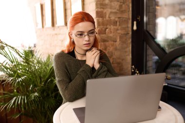 Kızıl saçlı genç bir kadın modern bir kafede oturuyor ve bilgisayarına odaklanıyor..
