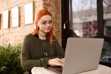 Bir kafede dizüstü bilgisayarı başında kahve yudumlayan ve mutlu hisseden genç bir kızıl..