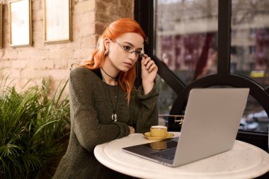 Kızıl saçlı güzel bir kadın modern bir kafede dizüstü bilgisayarıyla çalışıyor..