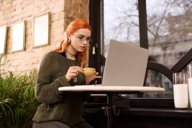 Odaklanmış, kızıl saçlı, laptopla çalışırken kahvenin tadını çıkaran genç bir kadın..