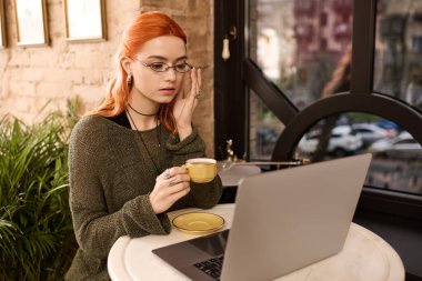 Güzel genç bir kadın, modern bir kafede dizüstü bilgisayarıyla çalışırken bir fincan kahveyle rahatlıyor..