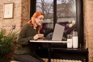 Parlak kızıl saçlı genç bir kadın kahveyi seviyor ve modaya uygun bir kafede dizüstü bilgisayarıyla çalışıyor..