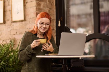 Şık bir kafede sıcak bir içecek tutarken kızıl saçlı neşeli genç bir kadın gülümsüyor..