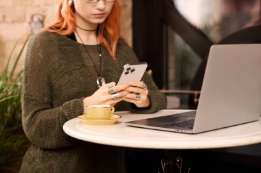 Canlı kızıl saçlı genç bir kadın şık bir kafede kahvesini yudumlarken telefonuyla meşgul oluyor..