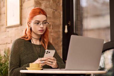 Kızıl saçlı genç bir kadın modern bir kafede rahat bir şekilde oturuyor, telefonuyla meşgul..
