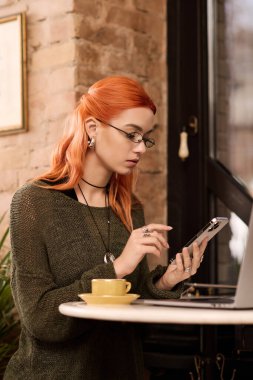 Kızıl saçlı güzel bir kadın, modern bir kafede telefonuna odaklanmış bir masada oturuyor..