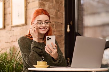 Samimi bir kafede telefonuna bakarken kızıl saçlı genç bir kadın gülümsüyor..