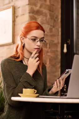 Canlı kızıl saçlı genç bir kadın düşünceli bir şekilde rahat bir kafede oturuyor.