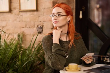 Kızıl saçlı bir kadın modern bir kafede dinleniyor, derin düşüncelere dalmışken içkisinin tadını çıkarıyor..