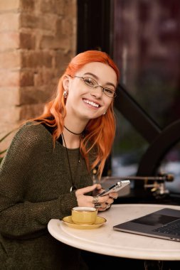 Kızıl saçlı neşeli genç bir kadın bir kafe masasında oturuyor ve telefonunu kullanırken gülümsüyor..