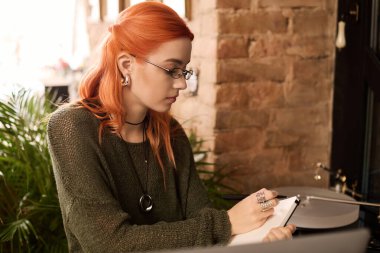 Modern bir kafede, kızıl saçlı genç bir kadın özenle çizimler yapar, yaratıcı düşüncelerine dalmıştır..
