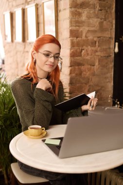 Kızıl saçlı genç bir kadın kafede eğleniyor, kahvesini yudumluyor ve yazıyor..