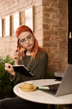 Kızıl saçlı genç bir kadın şık bir kafede defterine yazı yazarken kafa yoruyor..