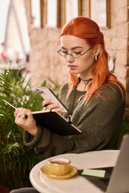 Kırmızı saçlı genç bir kadın, hareketli bir kafede kahvesini yudumlarken defterine yazıyor..