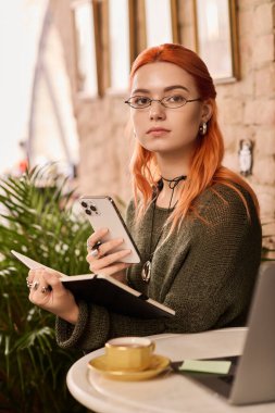 Kızıl saçlı güzel bir kadın modern bir kafede oturuyor, defterine ve telefonuna odaklanmış..