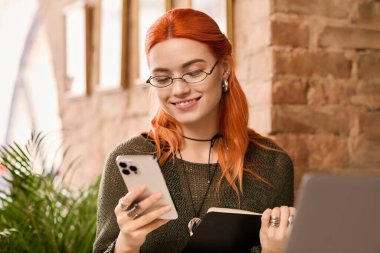 Kızıl saçlı genç bir kadın modern bir kafede oturuyor ve telefonuna gülümsüyor..