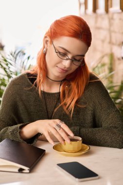 Kızıl saçlı genç bir kadın modern bir kafe ortamında kitap okurken sıcak bir içki içiyor..