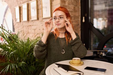 Kızıl saçlı genç bir kadın bir kafe masasında oturur, kahvesini yudumlar ve fikir üretir..