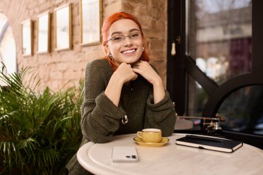 Modern bir kafede, kızıl saçlı genç bir kadın kahvesine gülümsüyor, rahat ve şık.