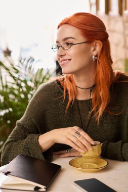 Canlı kızıl saçlı genç bir kadın elinde kahveyle şık bir kafede rahatlıyor..