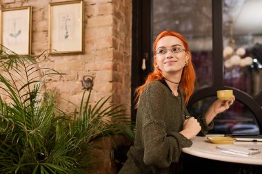 Çarpıcı kızıl saçlı genç bir kadın modern bir kafede rahatlıyor, içkisini yudumluyor ve gülümsüyor..