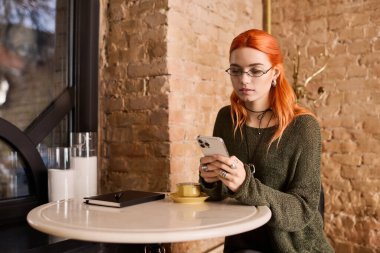 Canlı bir kızıl saçlı şık bir kafede oturur, kahvesini yudumlar ve akıllı telefonuna odaklanır..