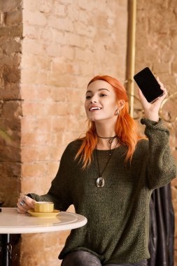 Parlak ve mutlu kızıl saçlı kadın, rahat bir kafede kahvesini yudumlarken telefonunu kaldırıyor..