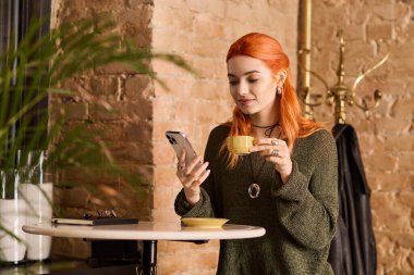 Canlı kızıl saçlı genç bir kadın modern bir kafede rahatlar ve içkisini yudumlar..