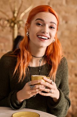 Genç bir kadın fincanından neşe saçarak yudumlarken parlak bir gülümseme kafeyi aydınlatıyor..