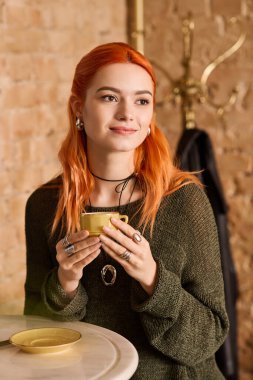 Çarpıcı kızıl saçlı genç bir kadın, modaya uygun bir kafede çayını yudumluyor..