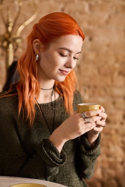 Kızıl saçlı neşeli genç bir kadın şık bir kafe ortamında sıcak bir fincan içiyor..