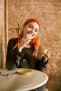 Kızıl saçlı neşeli genç bir kadın şık ve davetkar bir kafede otururken kahvesini yudumluyor..