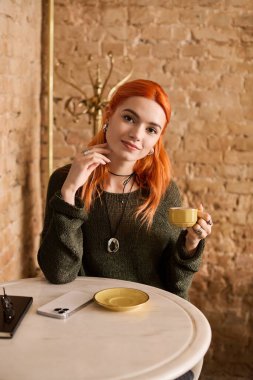 Kızıl saçlı genç bir kadın kahvesini yudumluyor, rahat, modern bir kafede dinleniyor..