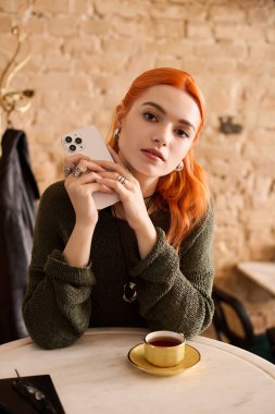 Güzel, genç bir kadın, kızıl saçlı, rahat bir kafede bir fincan kahve ve telefonuyla rahatlıyor..
