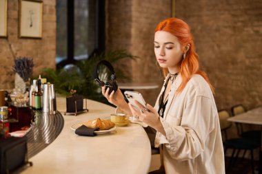 Kızıl saçlı genç bir kadın kahvesini yudumluyor ve bir kafede kruvasan yiyor..