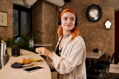 Kızıl saçlı genç bir kadın, rahat bir kafede kahvenin tadını çıkarıyor, en sevdiği melodilerde kaybolmuş..